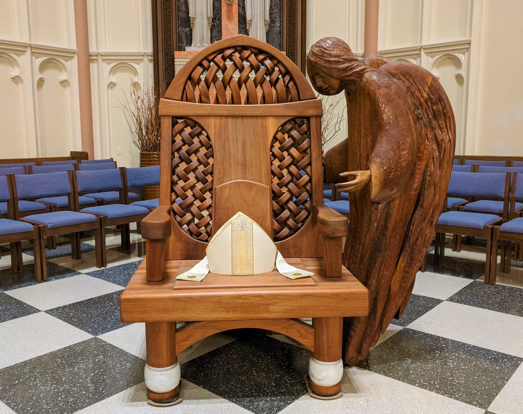 The wooden cathedra (bishop's seat). The cathedra has a wooden angel which appears to be whispering to the bishop when he is seated. On the cathedra is a white and gold mitre.