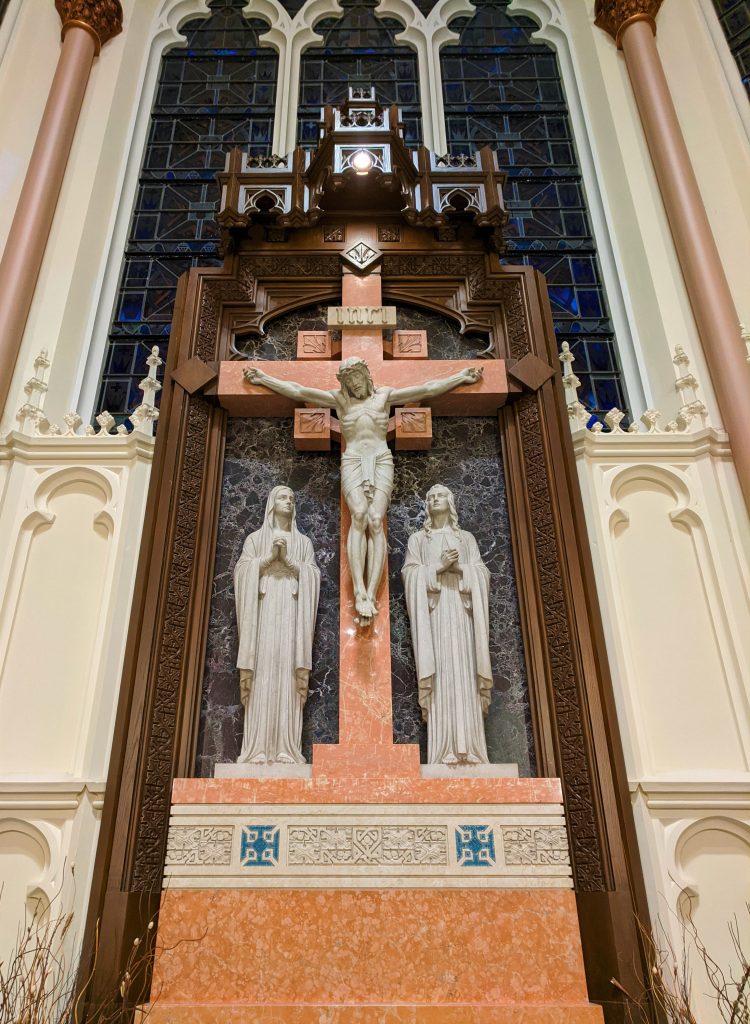 Large stone crucifix behind the altar. The cross is made of light orang stone. At the side of the the cross are the Blessed Mother and Saint John who, like Jesus, are made out of light grey stone.