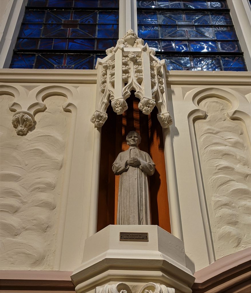 A small light grey stone statue of Saint John Bosco.