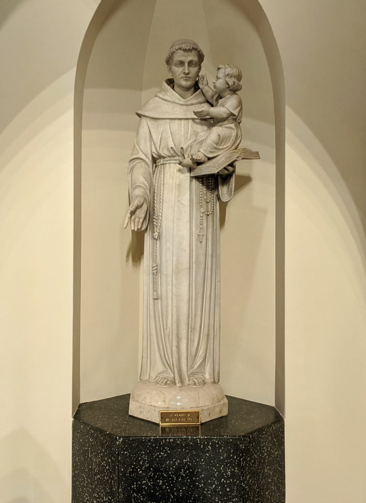 A light grey stone statue of Saint Anthony holding the child Jesus.